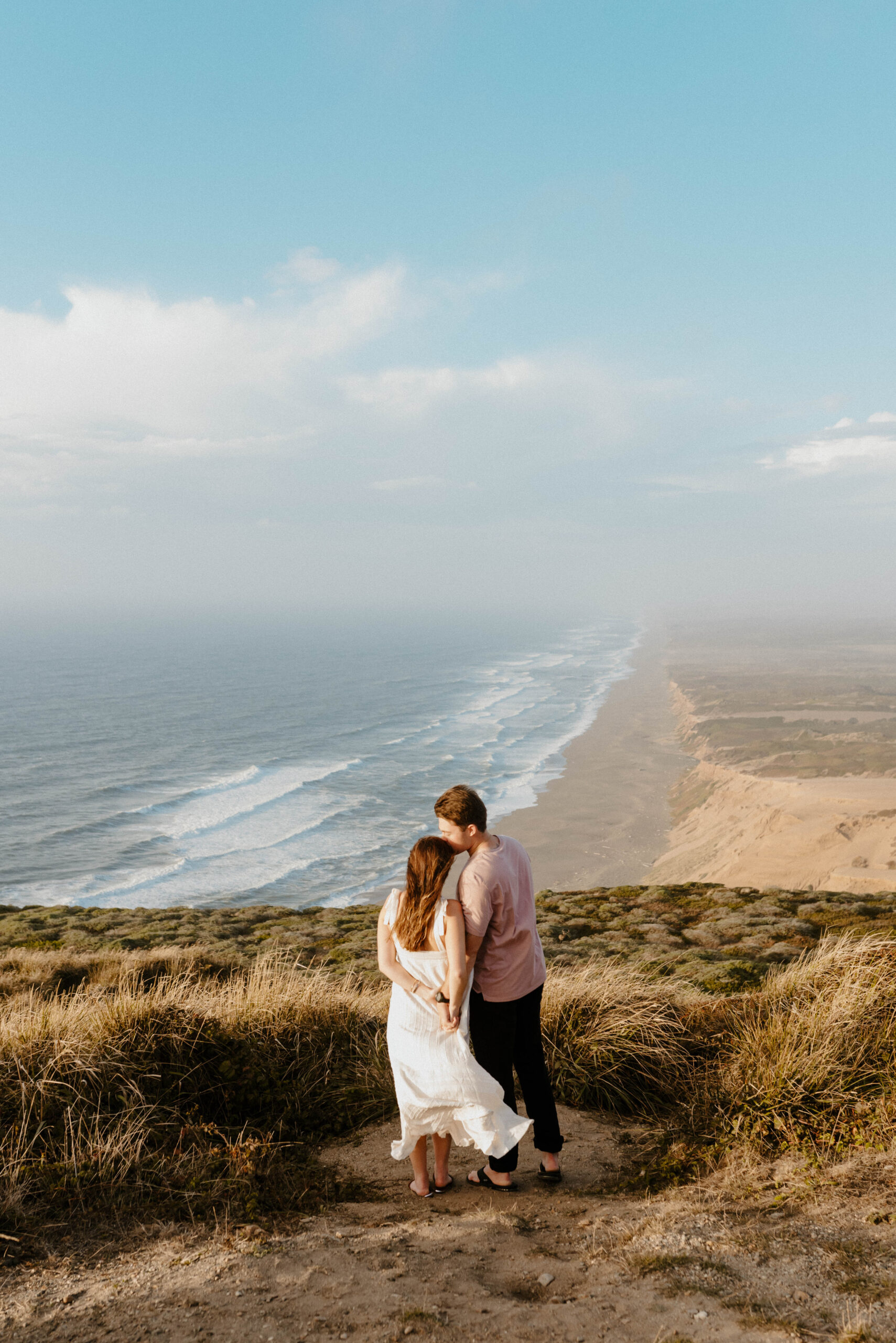 A couple - boy and girl - embracing while standing at the top of a cliff overlooking Point Reyes South Beach in California at golden hour